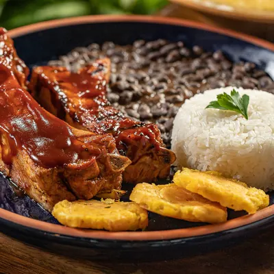 Costillas de cerdo glaseadas en salsa BBQ con menestra, arroz y patacones en plato de borde azul.