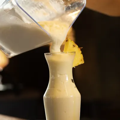 Garrafa de vidrio llena de bebida cremosa de pina con una rodaja de pina apoyada en el borde.