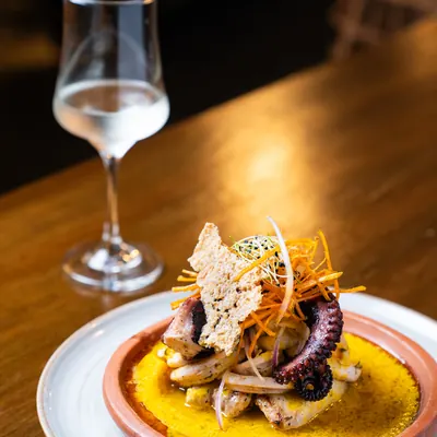 Ceviche a la parrilla servido en cazuela de barro, con pulpo, calamar y camarón sobre una crema amarilla, y una copa de vino blanco al fondo