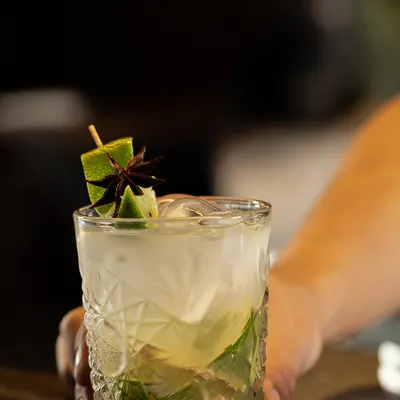 Coctel de lima en vaso de cristal tallado con brocheta de lima y anis estrellado, sobre barra de madera.
