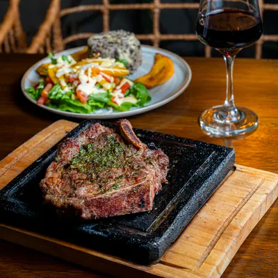 Corte T-bone con chimichurri sobre piedra caliente, ensalada al lado y una copa de vino tinto