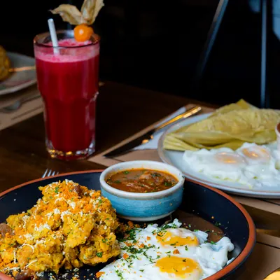 Tigrillo de verde majado con queso y dos huevos fritos en plato negro, con un bowl azul de salsa y un jugo rojo al lado