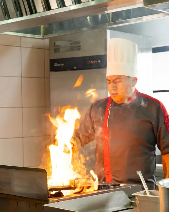 Cocinero con gorro alto flameando sobre la plancha en la cocina, con una llamarada encendida frente a él